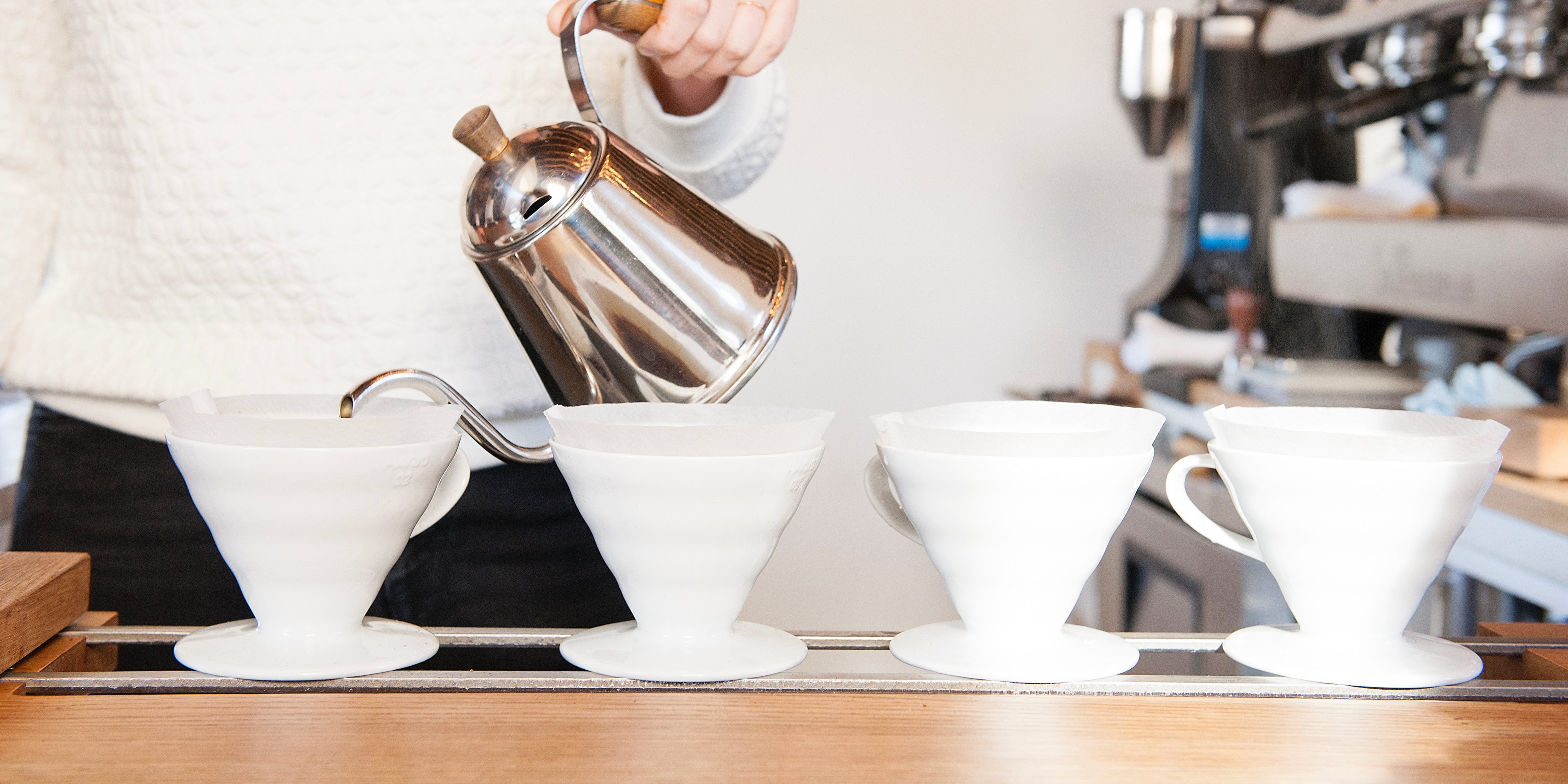 A person pours hot water from a metal kettle into one of four white ceramic pour-over coffee drippers lined up on a counter.