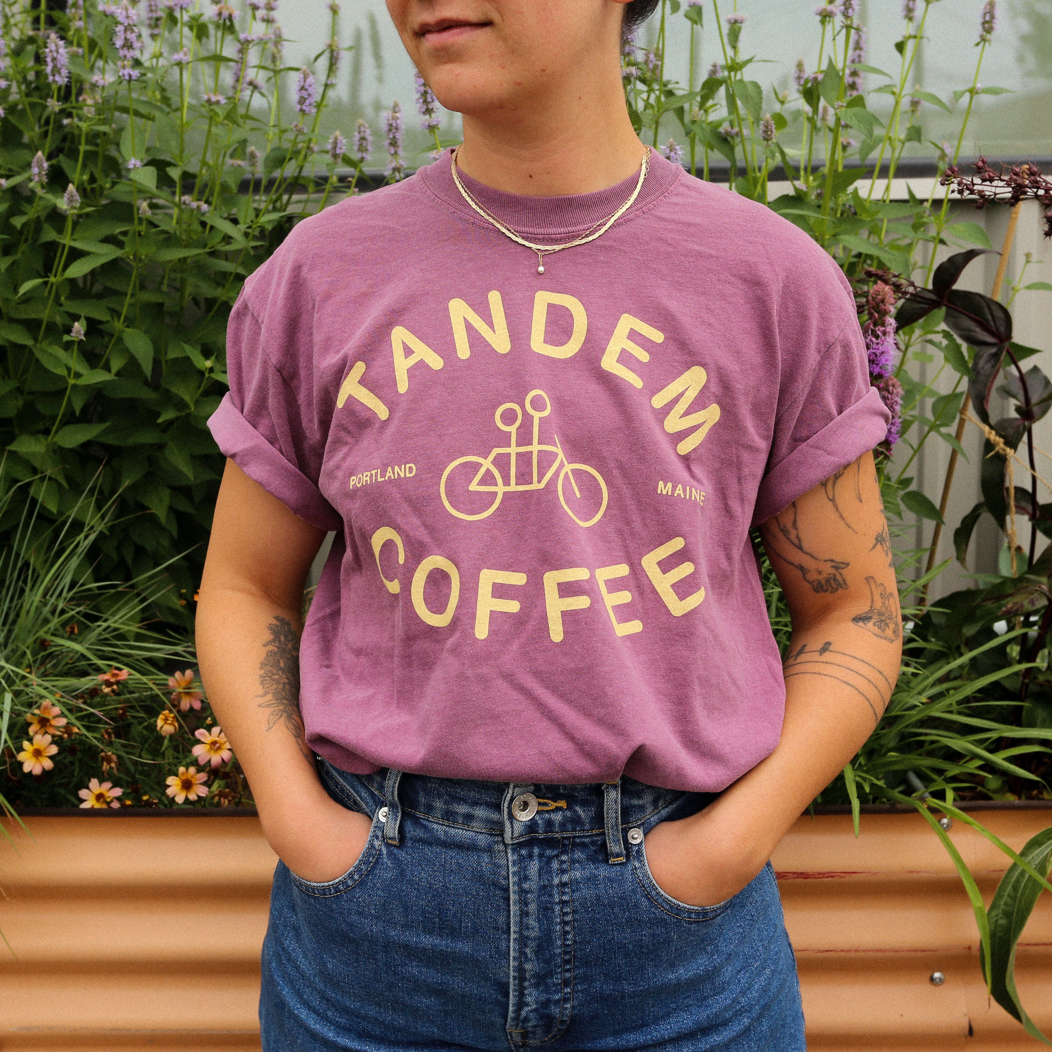 A person with short hair and visible arm tattoos stands outdoors among green plants and flowers, wearing the Comfort Colors Berry Logo Tee by Tandem Coffee Roasters. Made from 100% cotton, this purple tee features effortless style with its signature logo design.