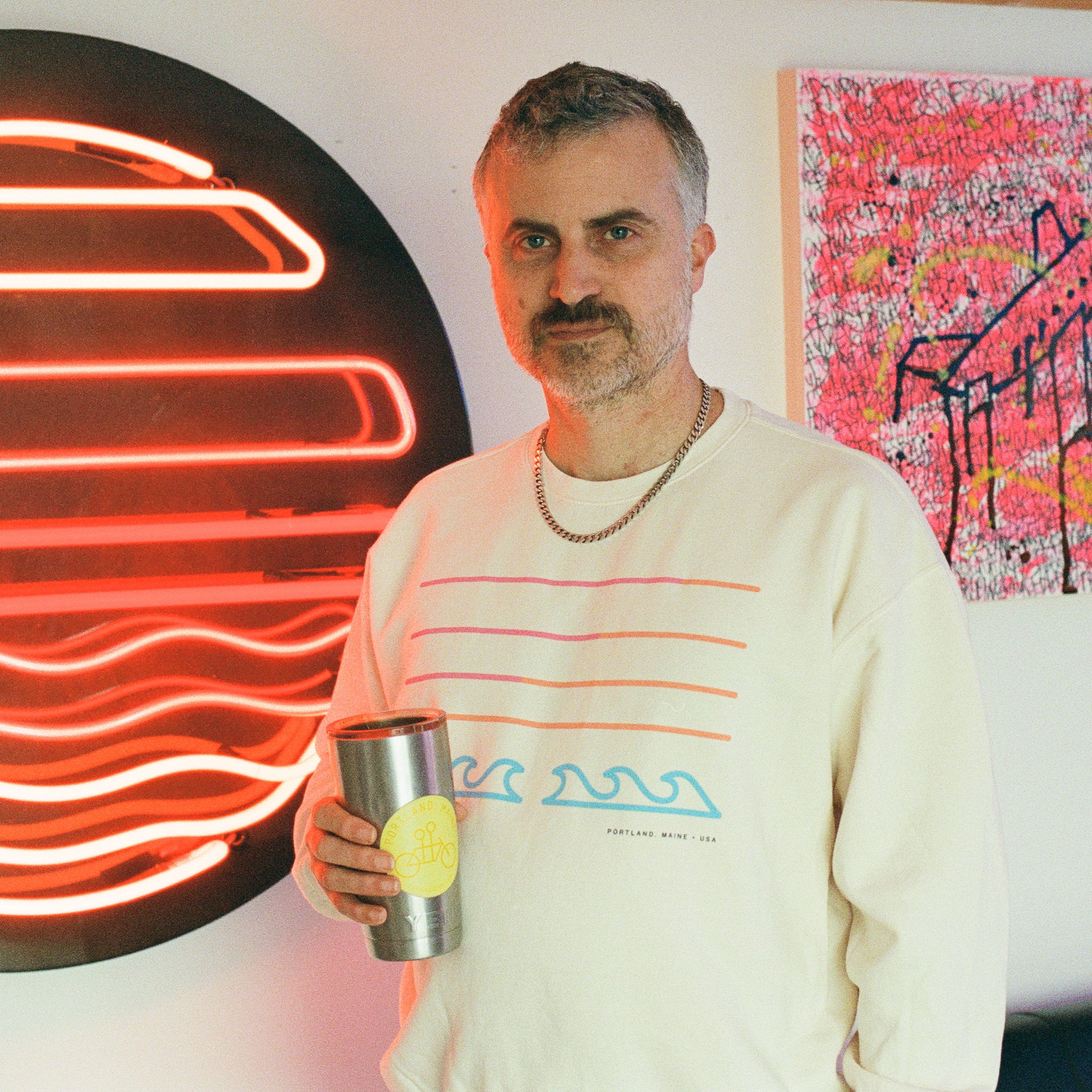 A man with short gray hair and a mustache stands indoors, holding a metal tumbler. He wears a Tandem Coffee Roasters Filthy Sunset Pullover. Behind him are a neon red wave sign and colorful Filthy Sunset Neon artwork on the wall.