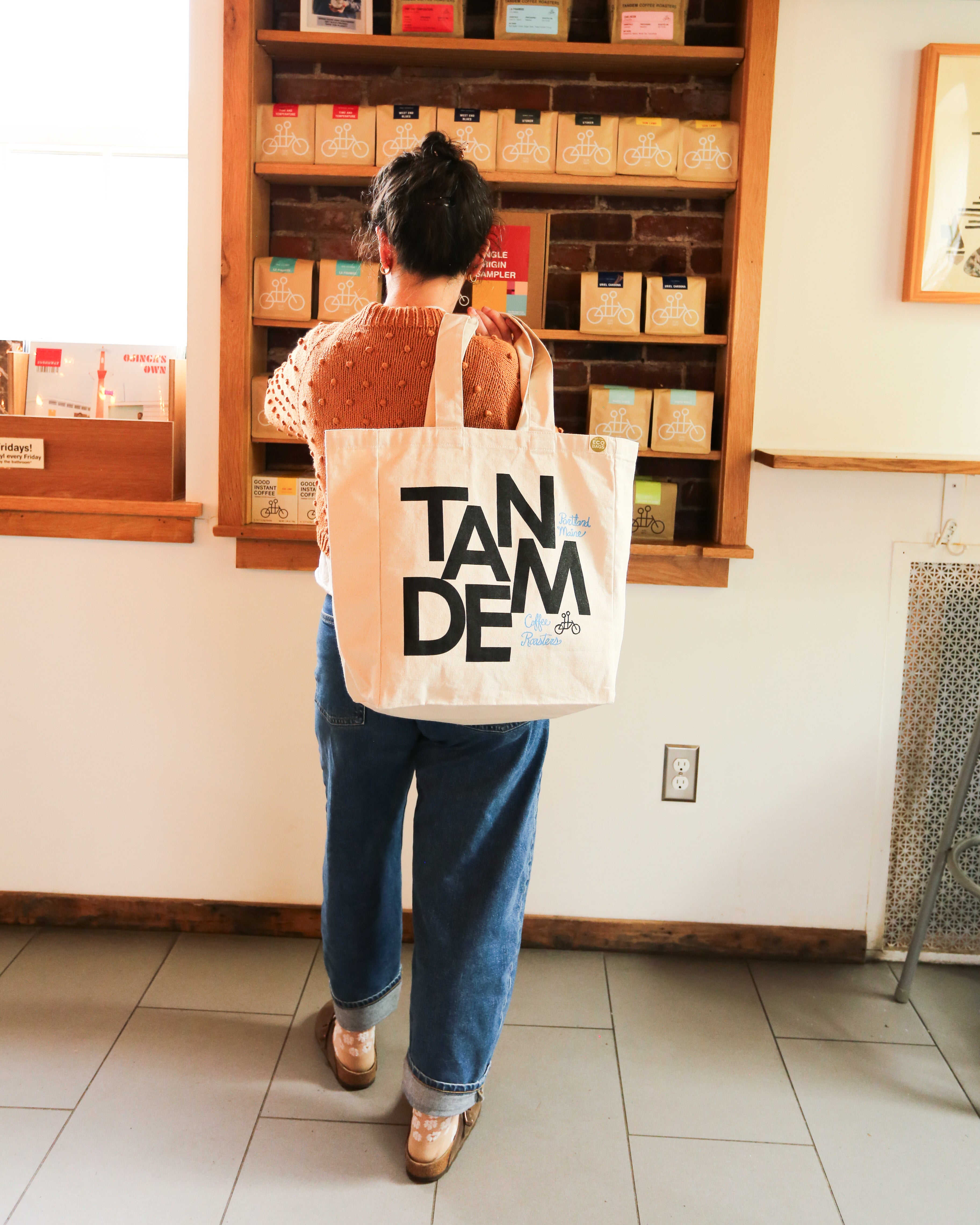 A person with The MONK Tote by Tandem Coffee Roasters stands before a wooden display of small boxes, facing away in a cozy shop, showcasing the use of reusable shopping bags.