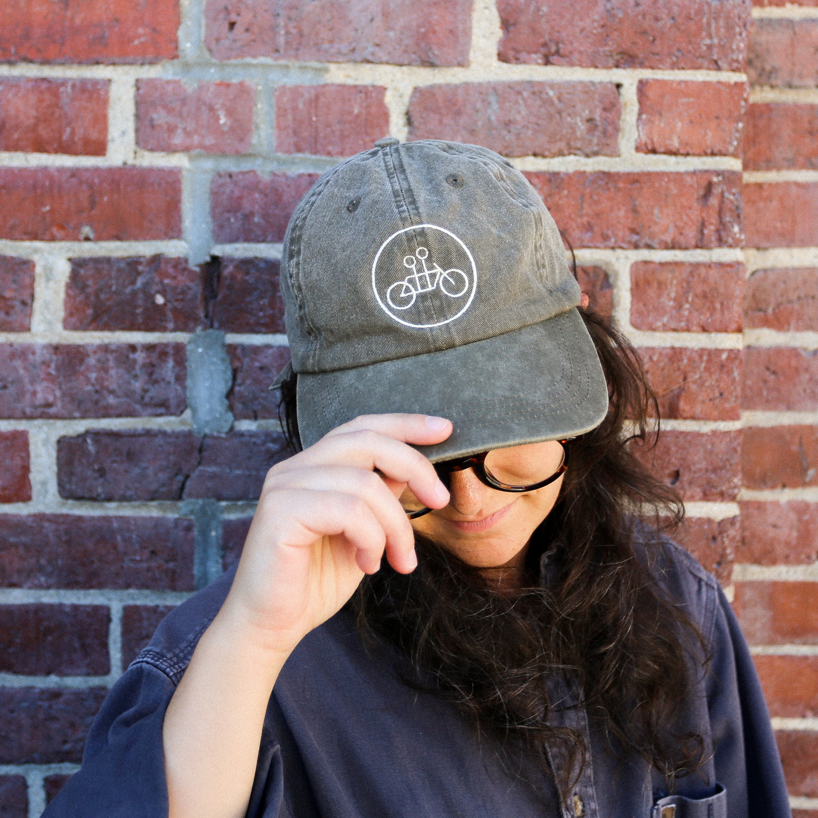 A person wearing the Tandem Coffee Roasters Java Circle Logo Dad Hat in green, tilting the cap downward while standing against a brick wall. Their long hair is loose, and they are wearing glasses and a blue shirt.
