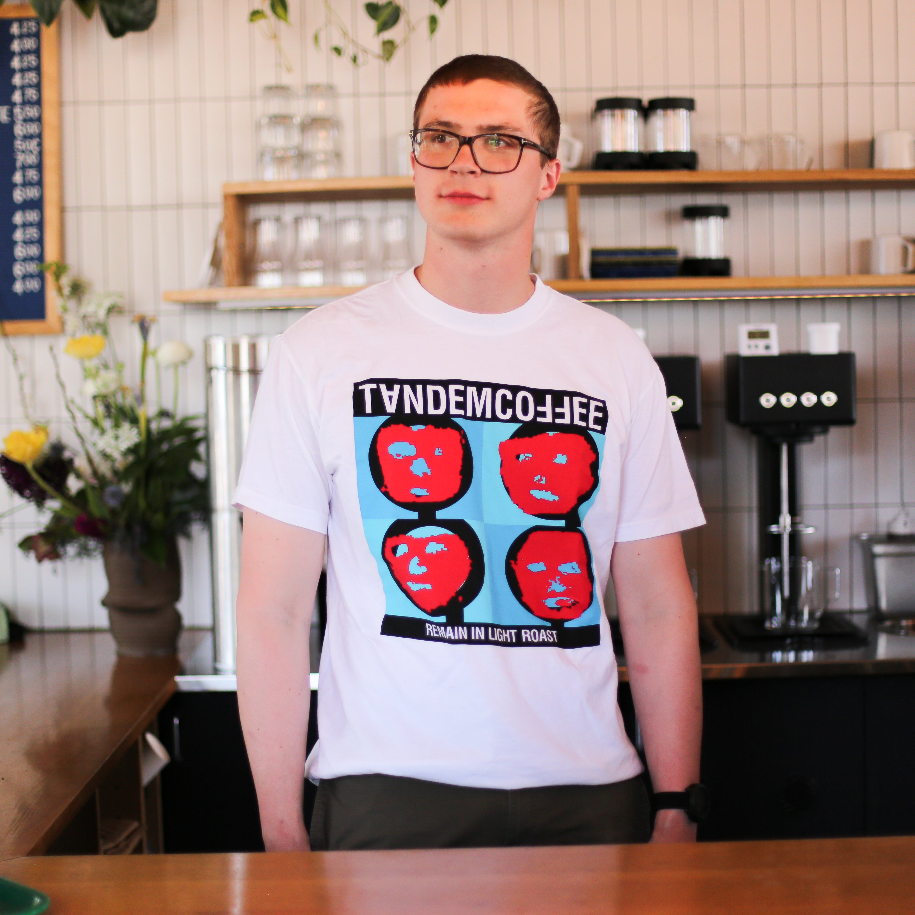 A man with glasses stands in a coffee shop wearing a white Remain In Light Roast T-Shirt by Tandem Coffee Roasters, printed in Maine with four red faces on a blue background and the text "Tandem Coffee" and "Remain in Light Roast.