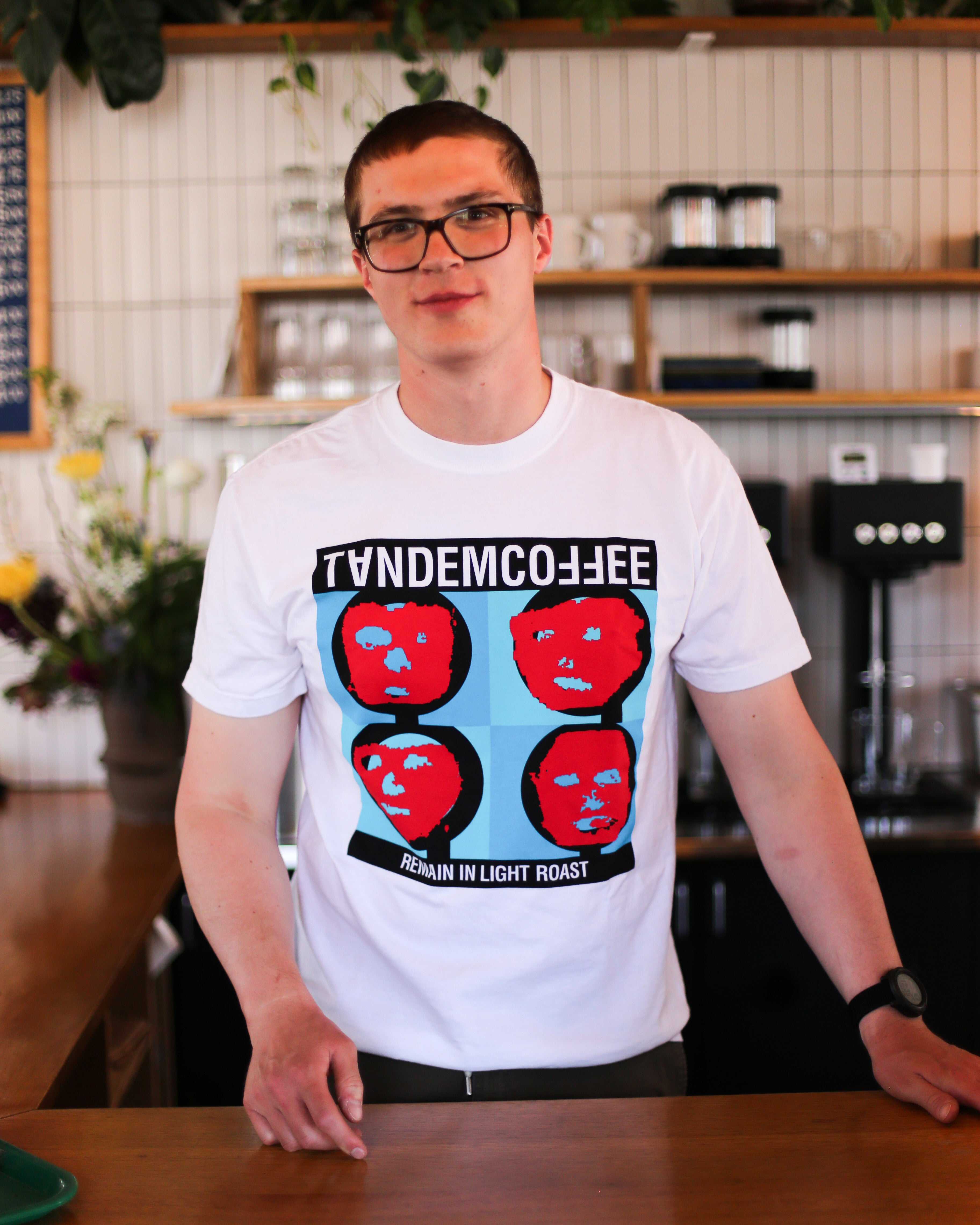 A man in glasses and a white Tandem Coffee Roasters Remain In Light Roast T-Shirt, featuring a pop-art design of four red faces and printed in Maine, stands behind a wooden counter in a coffee shop with flowers and shelves behind him.