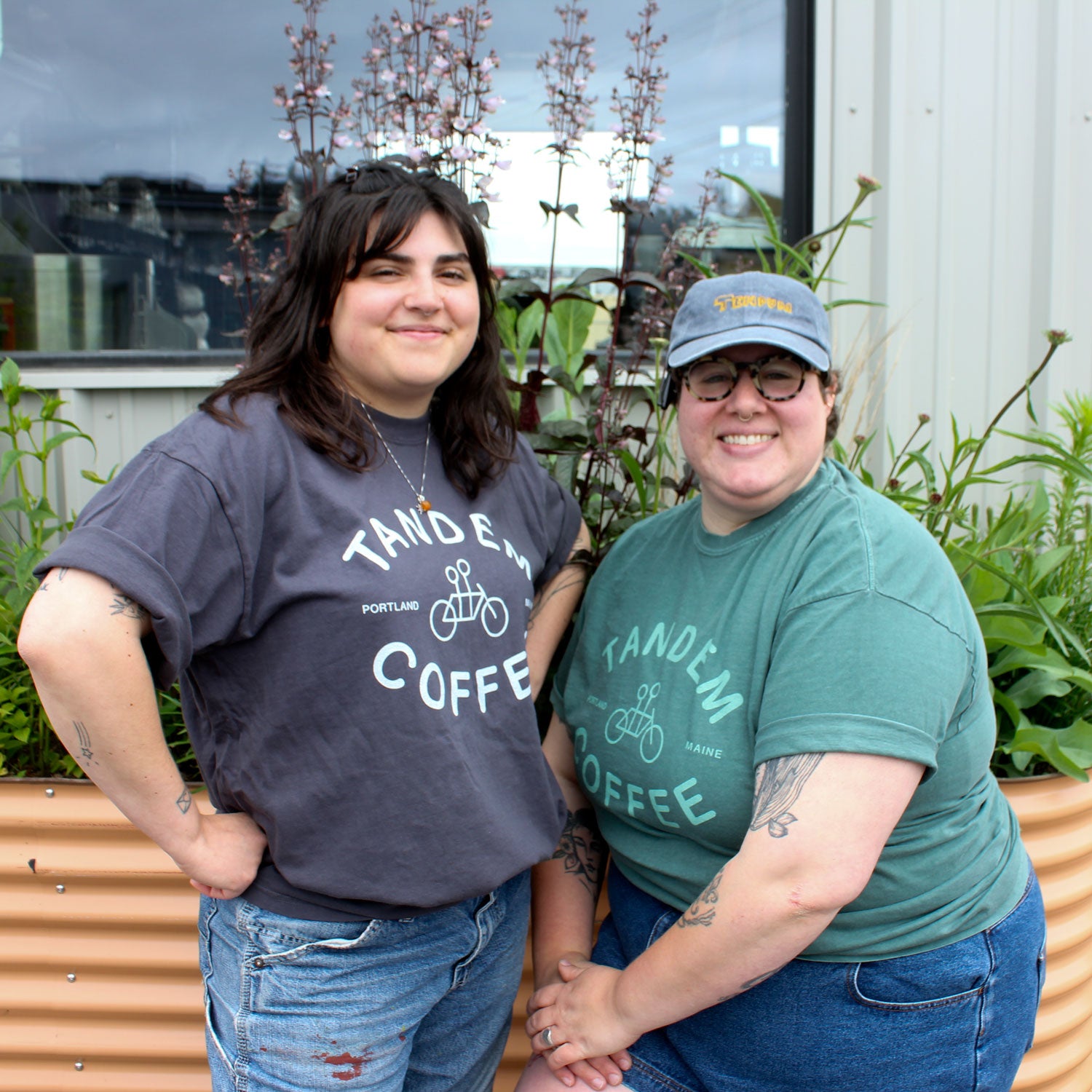 Two people, both wearing Tandem Coffee Roasters Graphite Logo Tee, smile outdoors in front of plants. Each wears glasses and jeans; one has a cap. Tattoos are visible on their arms.