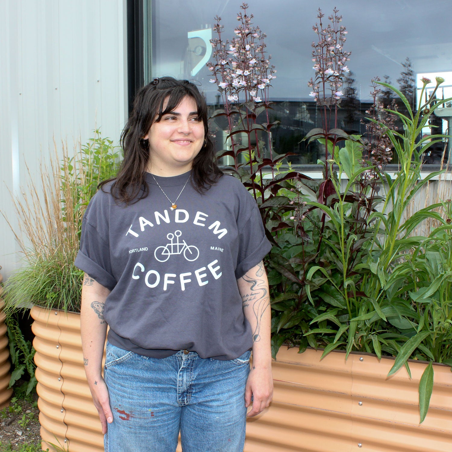 A person with dark hair and tattoos wears a Tandem Coffee Roasters Graphite Logo Tee and blue jeans, smiling while standing in front of a raised garden bed filled with green and purple plants.