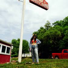 A woman in casual clothes stands on grass near a tall Waxahatchee - Tigers Blood signpost from Tandem Coffee Roasters outside a red building, with trees and a red pickup truck in the background under a cloudy sky.