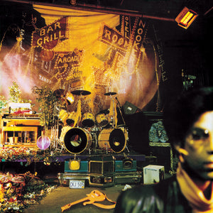 A cluttered stage with a drum set, keyboard, flowers, and traffic signs evokes the vibrant chaos of Tandem Coffee Roasters’ Prince - Sign o' the Times, as a blurred figure appears in the right foreground.