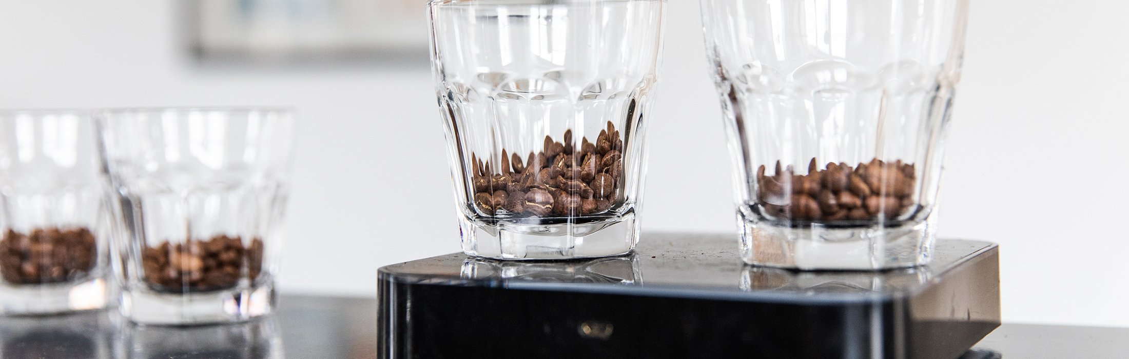 Close-up of clear glass cups with coffee beans inside, some on a black digital scale and others in the background on a white surface.