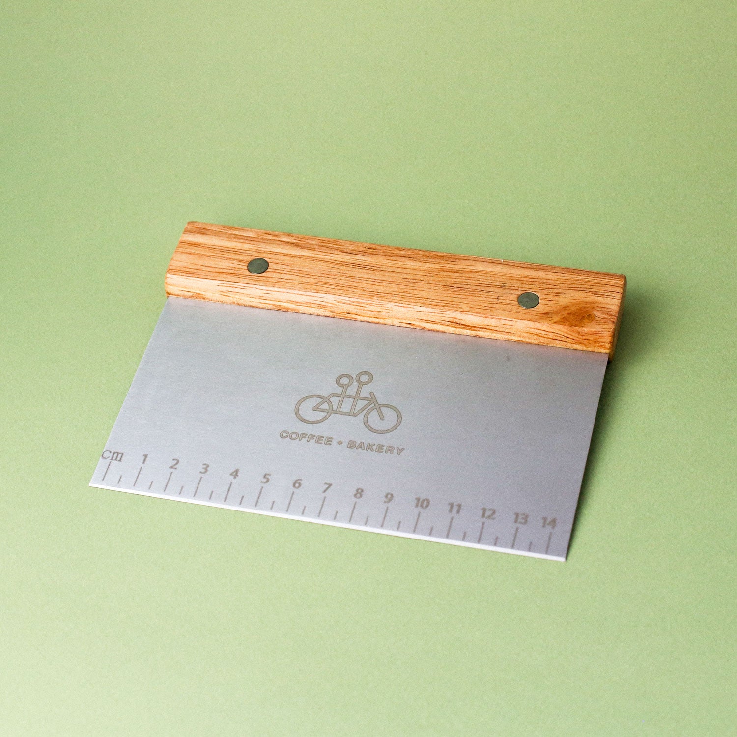 The Tandem Bench Scraper by Tandem Coffee Roasters, with a wooden handle and engraved ruler, rests on a light green surface. Its metal blade features a bicycle and "COFFEE & BAKERY," ideal for precise dough handling.