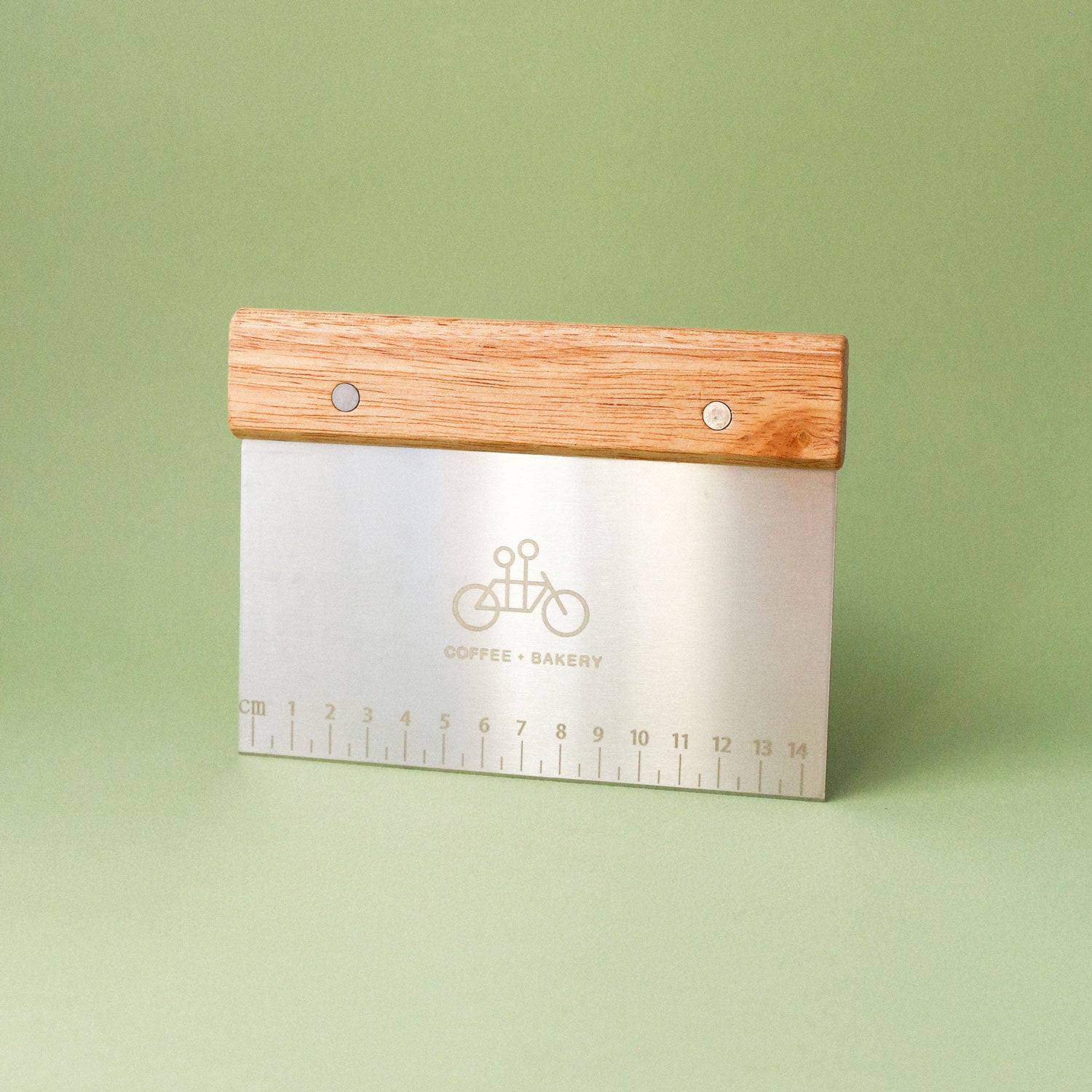 The Tandem Bench Scraper by Tandem Coffee Roasters features a wooden handle, a metal blade engraved with a bicycle and COFFEE + BAKERY, and a built-in centimeter ruler along the bottom edge. It stands upright against a green background.