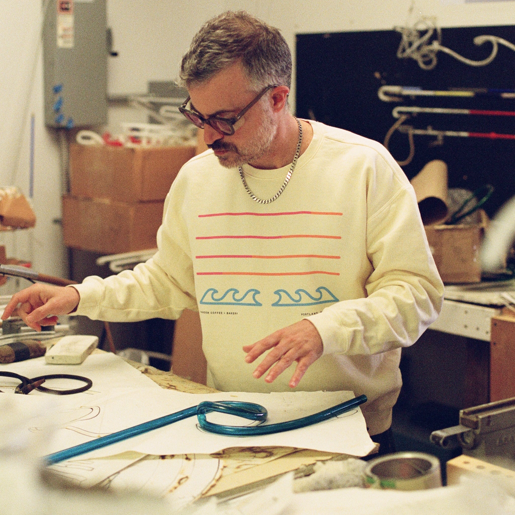 A man in glasses wearing a Tandem Coffee Roasters Filthy Sunset Pullover with wave designs works at a cluttered table, measuring or inspecting a neon blue strip amid papers and tools in his workshop.