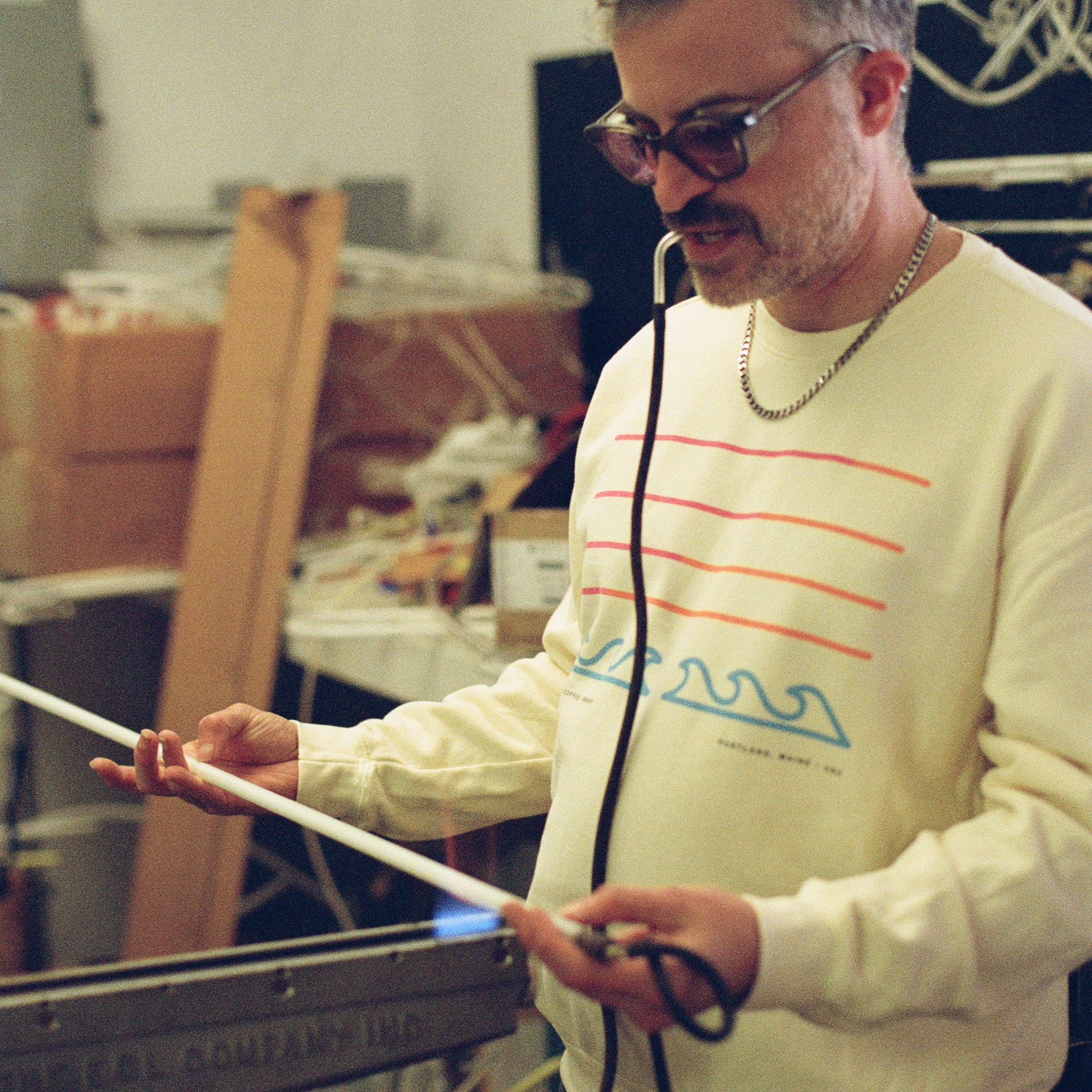 A man wearing sunglasses and a Filthy Sunset Pullover by Tandem Coffee Roasters holds a long, thin rod and grips a cable in his mouth, standing in a cluttered room with equipment and boxes in the background.