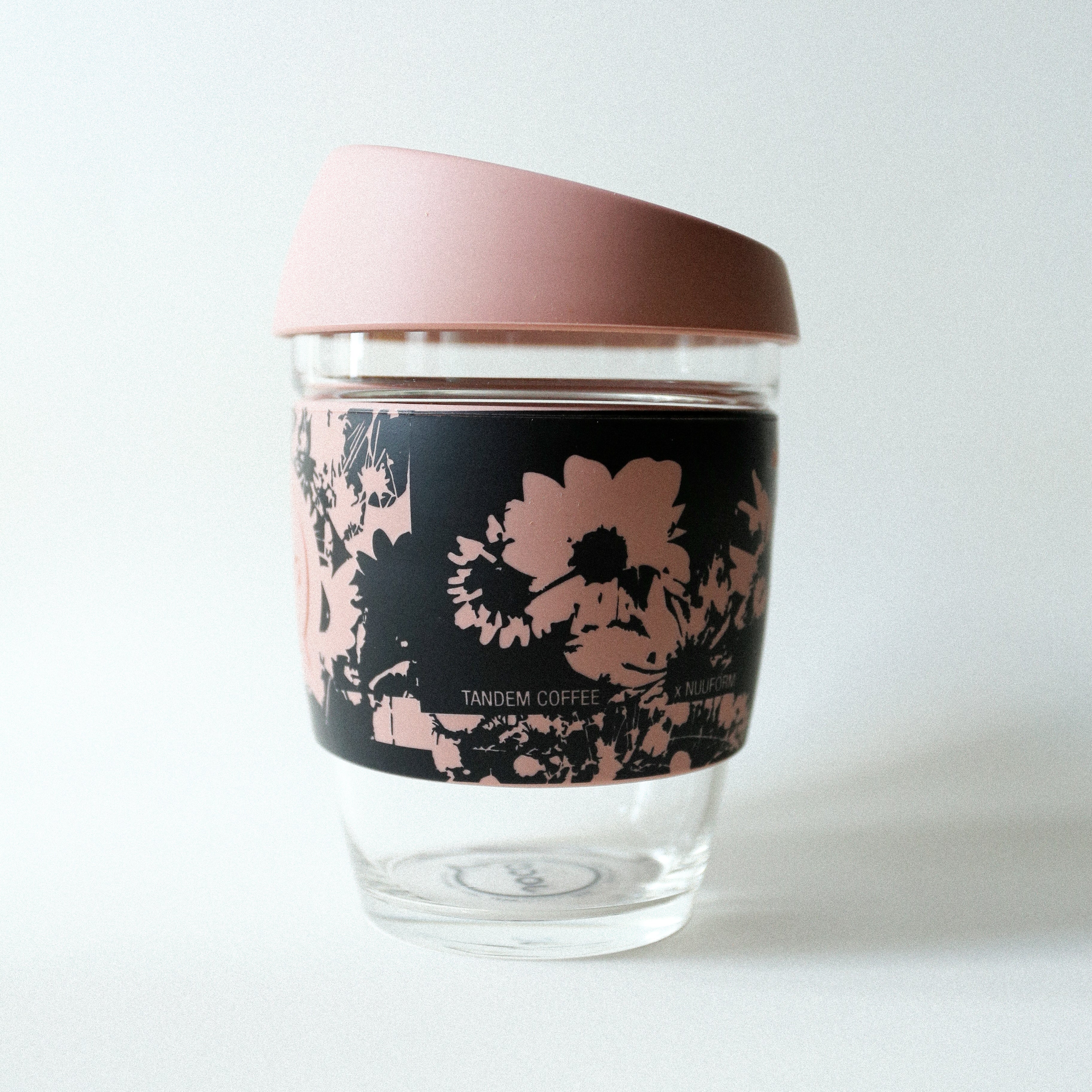 The Tandem x NUUFORM Joco Cup by Tandem Coffee Roasters is a plastic-free, reusable borosilicate glass coffee cup with a pink lid and a black sleeve decorated with pink floral patterns, shown on a white background.