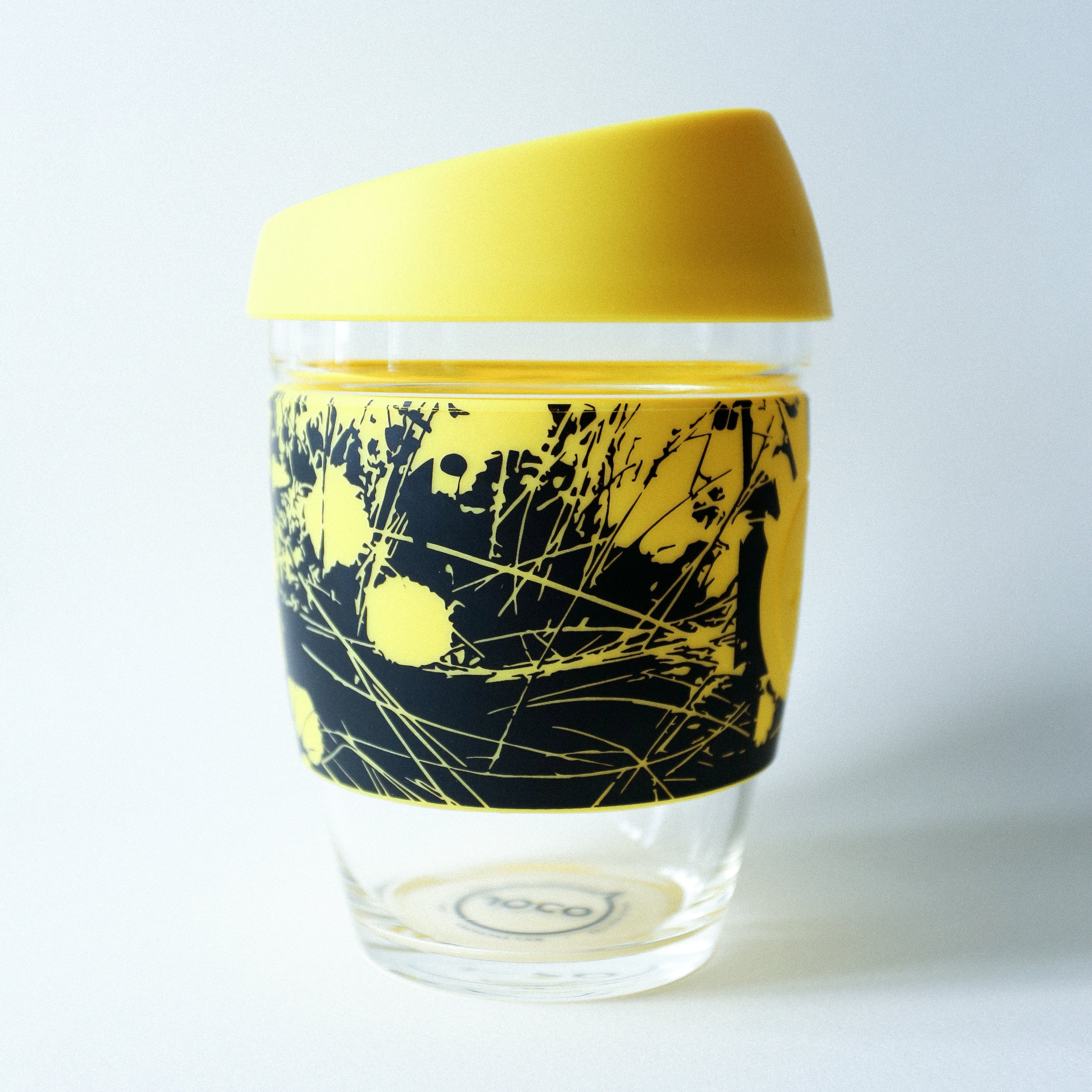 The Tandem x NUUFORM Joco Cup by Tandem Coffee Roasters is a reusable borosilicate glass cup with a yellow silicone lid and a yellow-and-black grass-patterned sleeve, crafted from an exclusive artist collaboration.