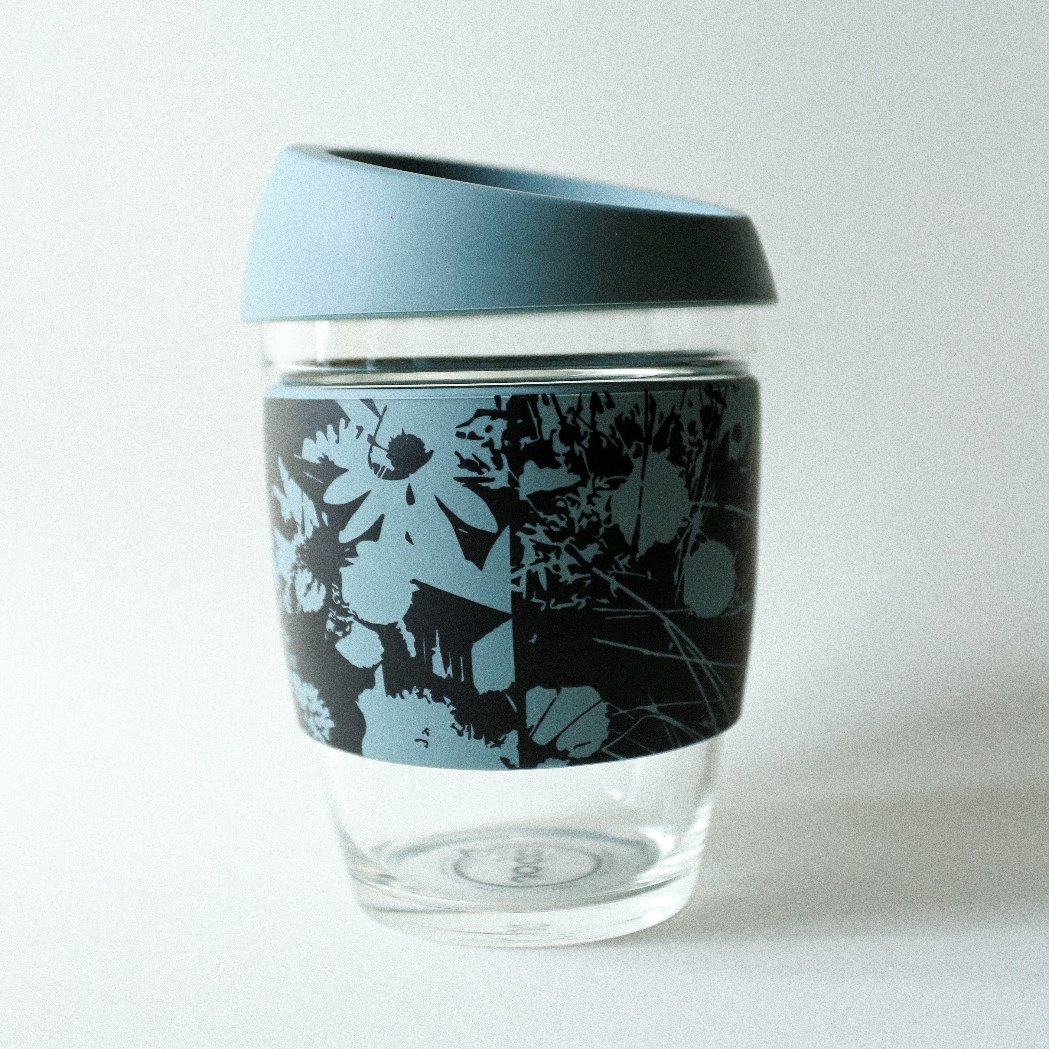 The Tandem x NUUFORM Joco Cup by Tandem Coffee Roasters is a plastic-free, reusable borosilicate glass coffee cup with a light blue lid and a black silicone sleeve featuring a light blue floral pattern on white.