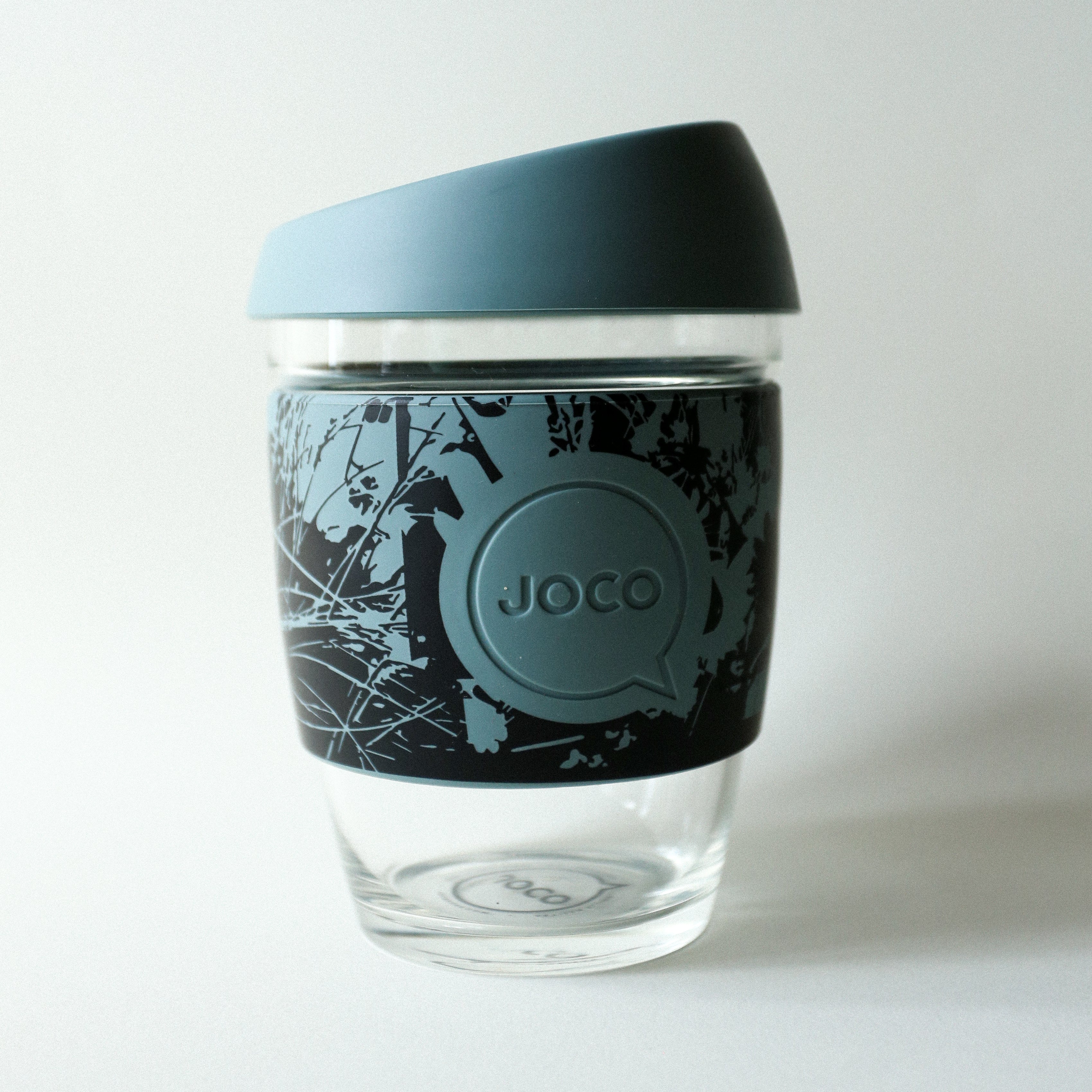 The Tandem x NUUFORM Joco Cup by Tandem Coffee Roasters is a plastic-free, reusable borosilicate glass cup with a blue silicone lid and nature-inspired sleeve, embossed with the JOCO logo. An elegant artist collaboration piece.