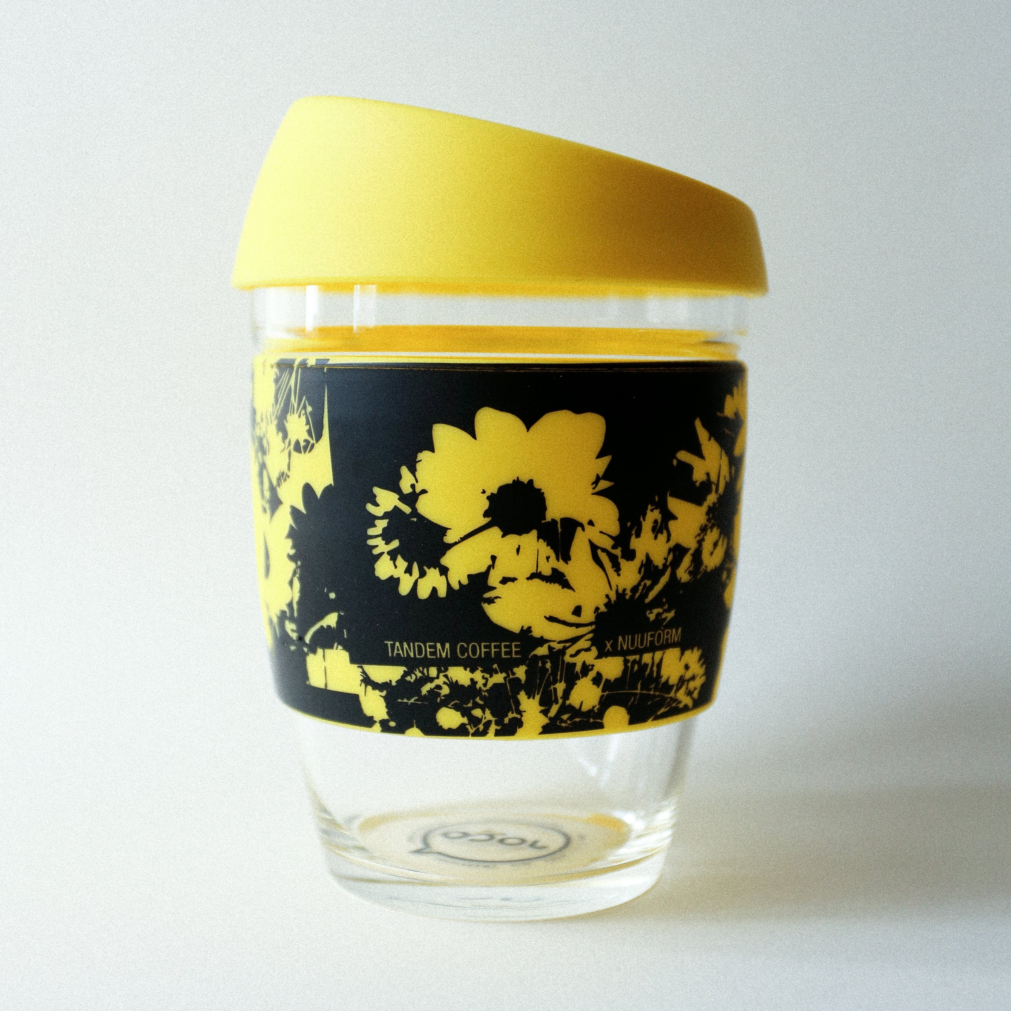 The Tandem x NUUFORM Joco Cup by Tandem Coffee Roasters is a plastic-free, reusable borosilicate glass cup with a yellow lid and black sleeve featuring bright yellow floral art from an artist collab, and "Tandem Coffee x MiiR" printed on the sleeve.