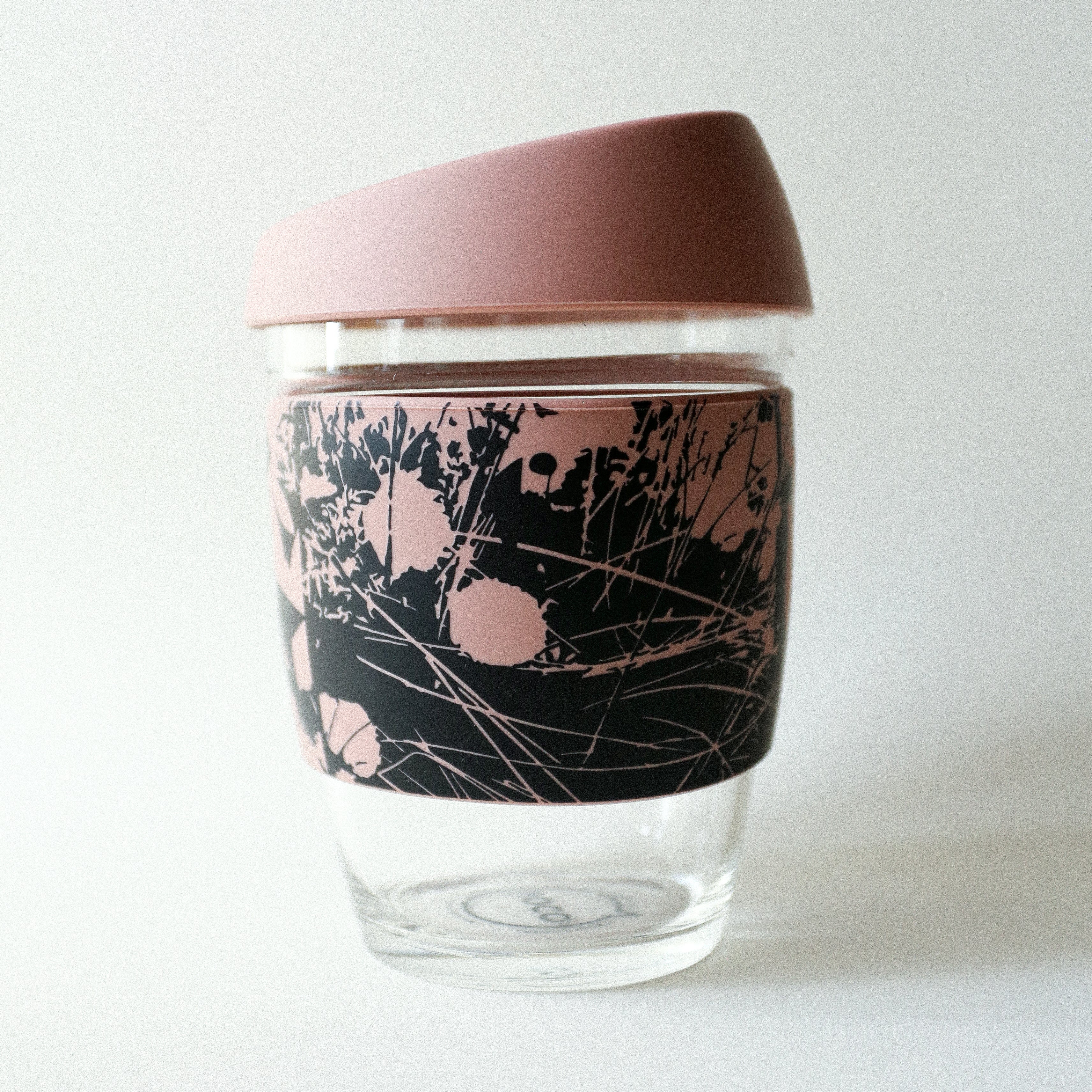The Tandem x NUUFORM Joco Cup by Tandem Coffee Roasters is a borosilicate glass cup with a pink lid and matching pink/black abstract plant sleeve, created in collaboration with an artist, shown against a plain white background.