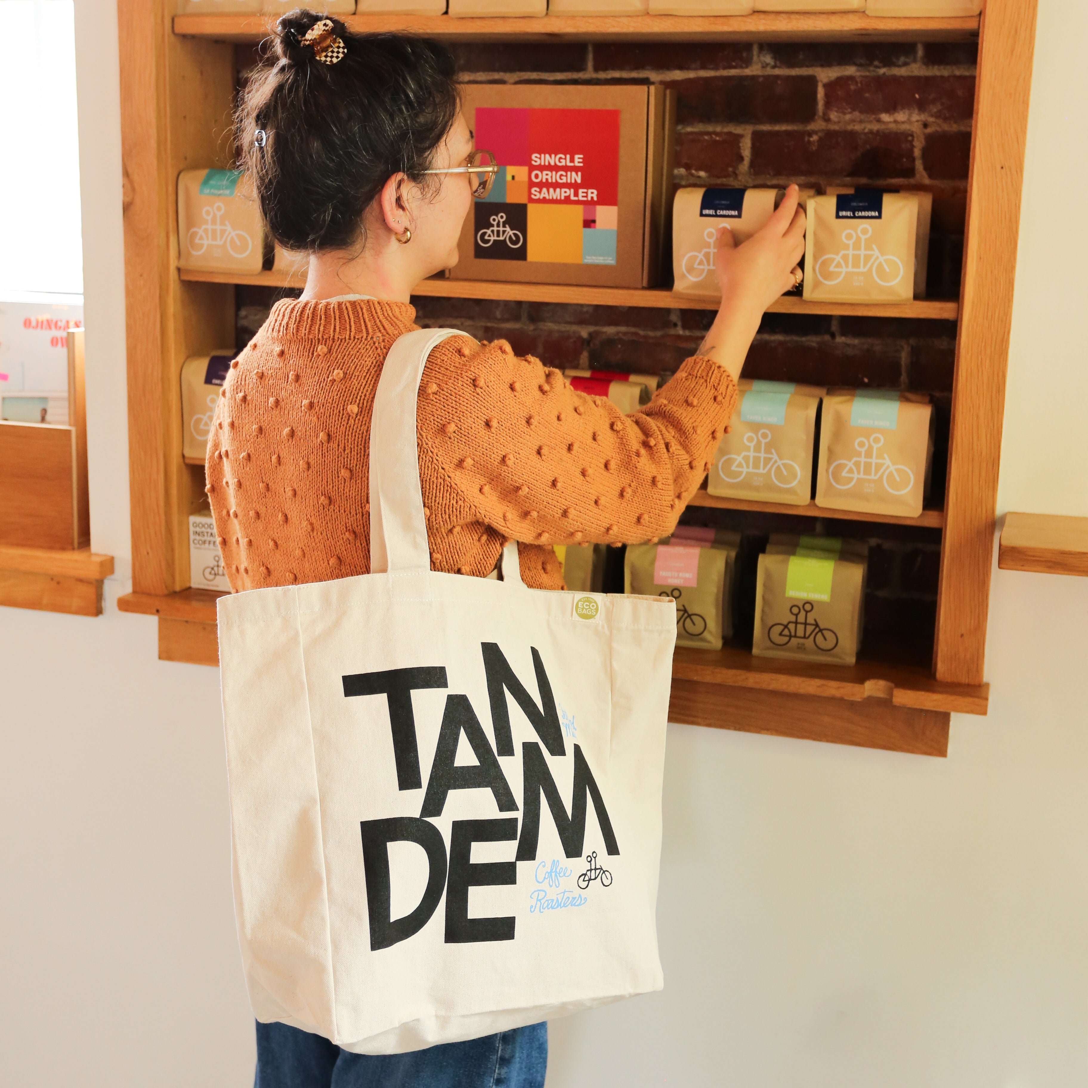 A person with glasses, a bun, and an orange textured sweater browses packaged coffee on wooden shelves in a cozy shop, carrying The MONK Tote by Tandem Coffee Roasters—a stylish, reusable beige canvas bag.