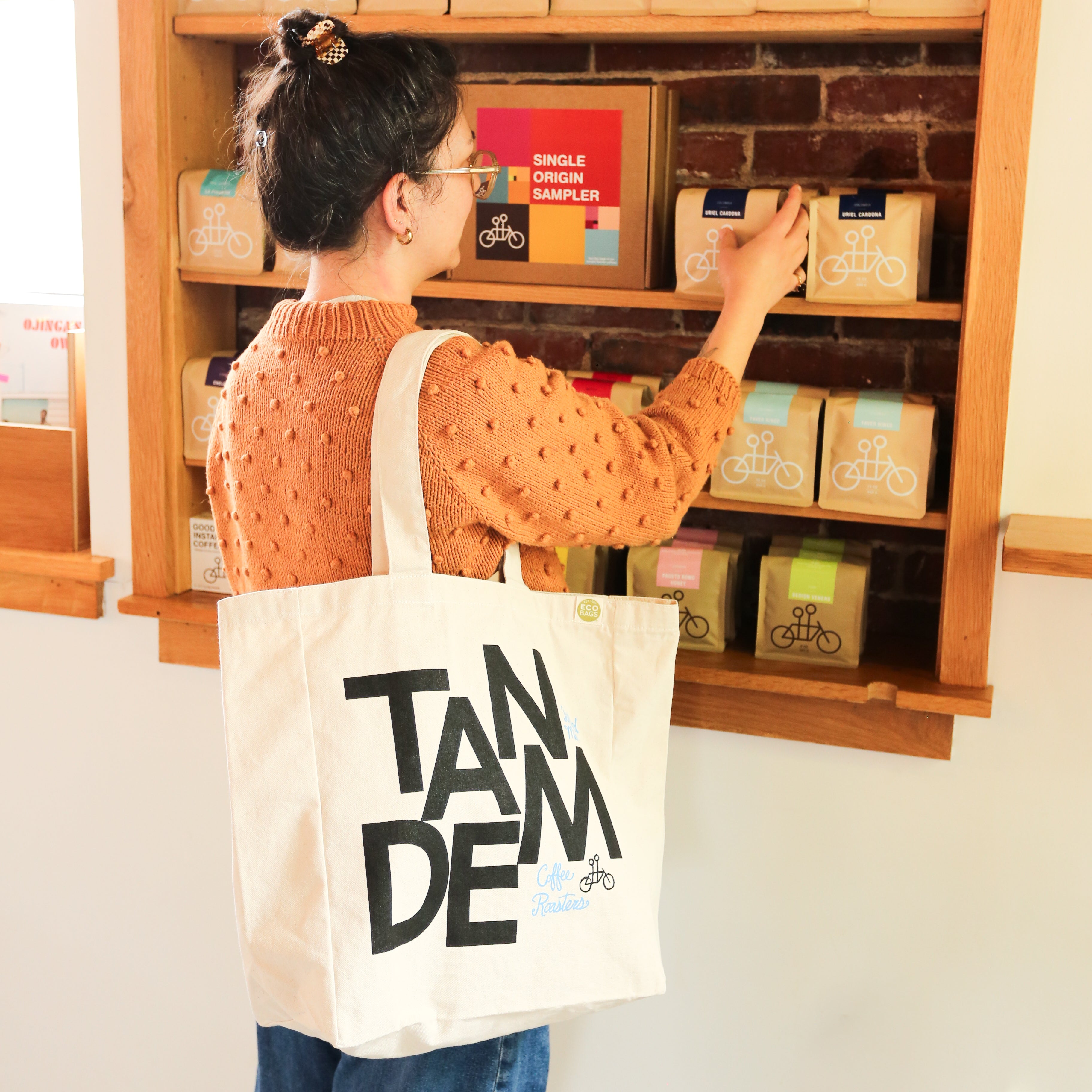 A person wearing glasses and an orange sweater browses coffee bags on a shelf, carrying a large MONK Tote by Tandem Coffee Roasters, with "Tandem" boldly printed on the canvas.