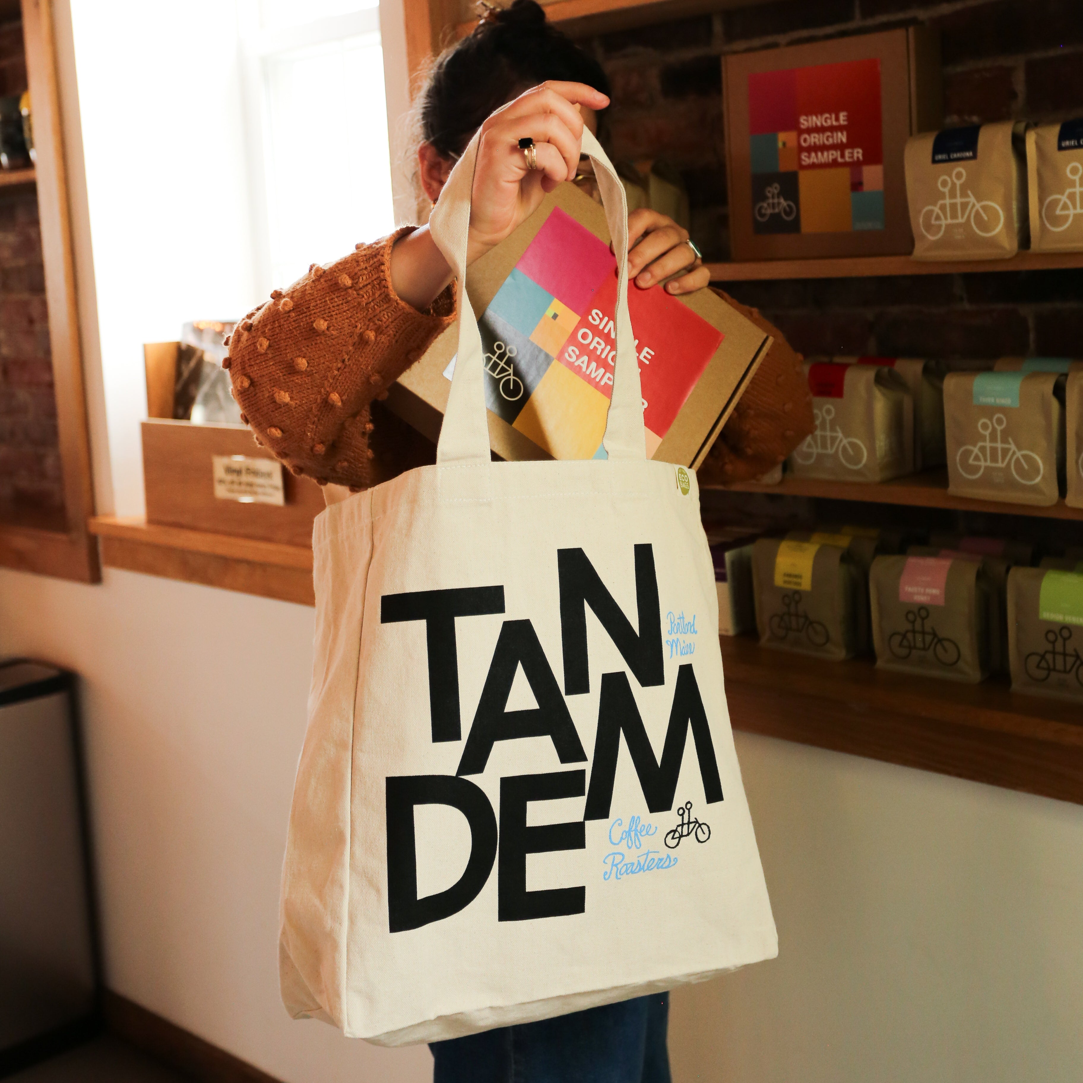 A person puts a Single Origin Sampler box into The MONK Tote by Tandem Coffee Roasters while standing in a shop with coffee products on display.