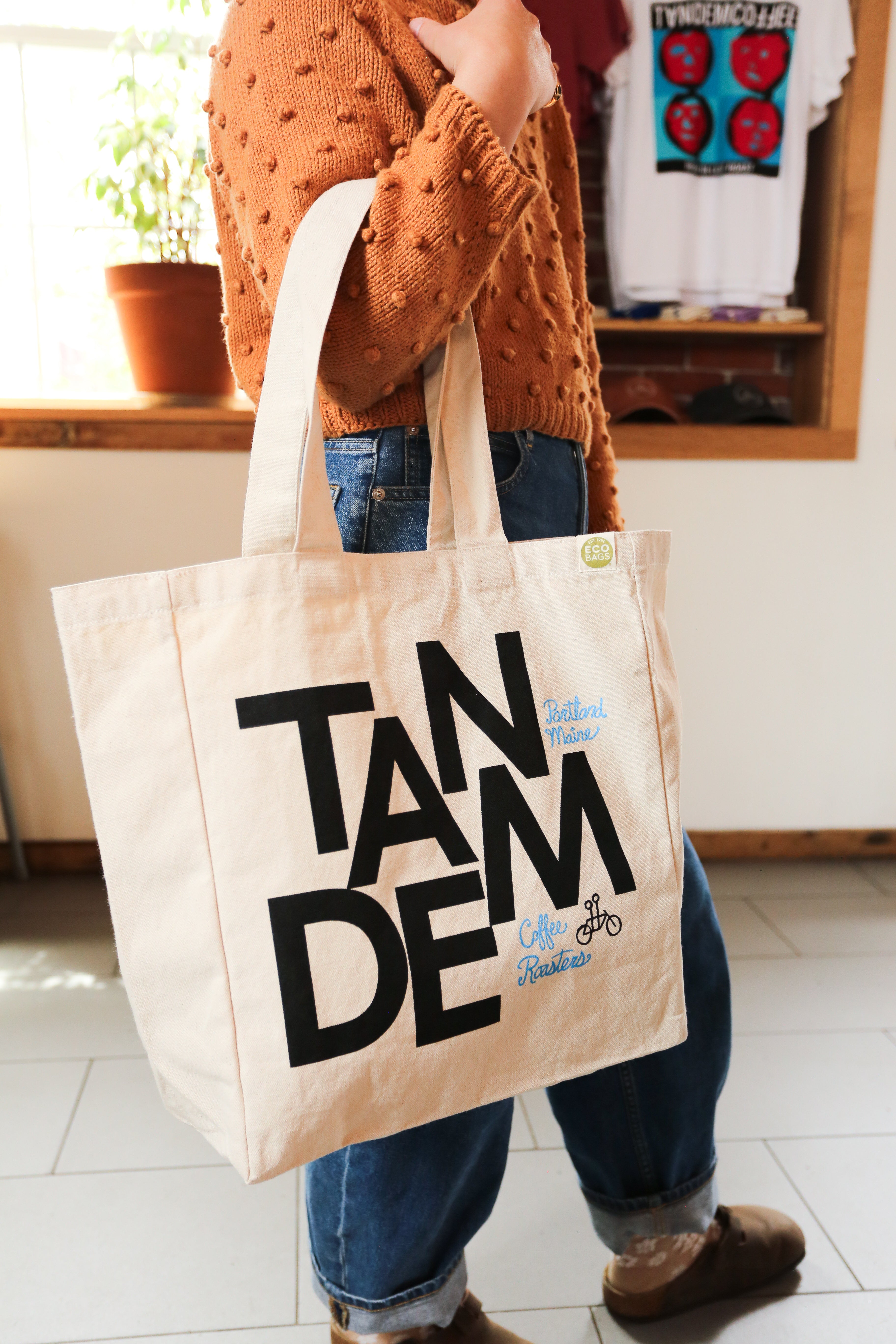 A person wearing a textured brown sweater and blue jeans holds The MONK Tote by Tandem Coffee Roasters, a large canvas bag with bold black "TANDEM" text and smaller blue "Coffee Roasters," perfect as a reusable shopping bag.