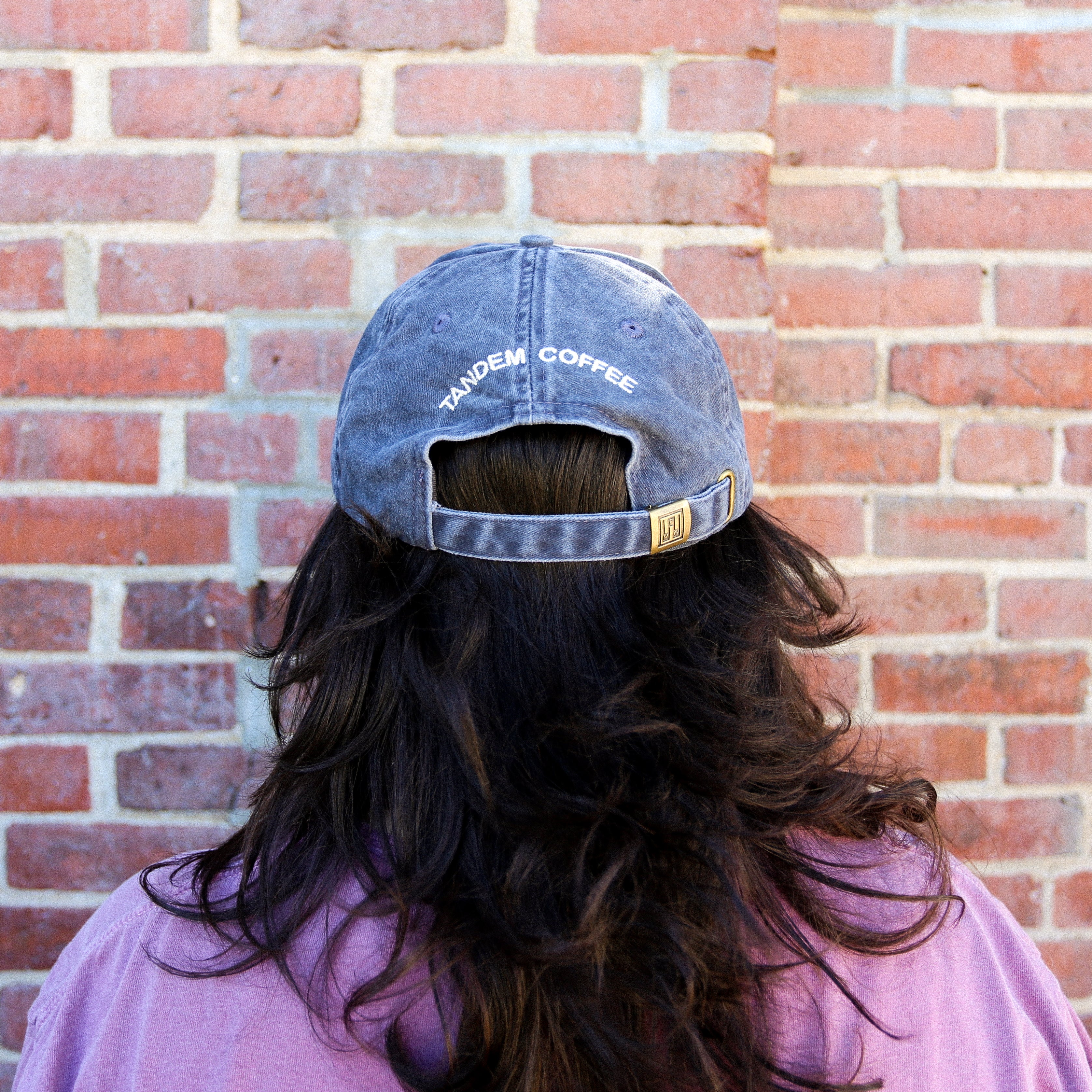 Person with long dark hair wearing a purple shirt and a Tandem Coffee Roasters Slate Blue Circle Logo Dad Hat stands facing a red brick wall, seen from behind.