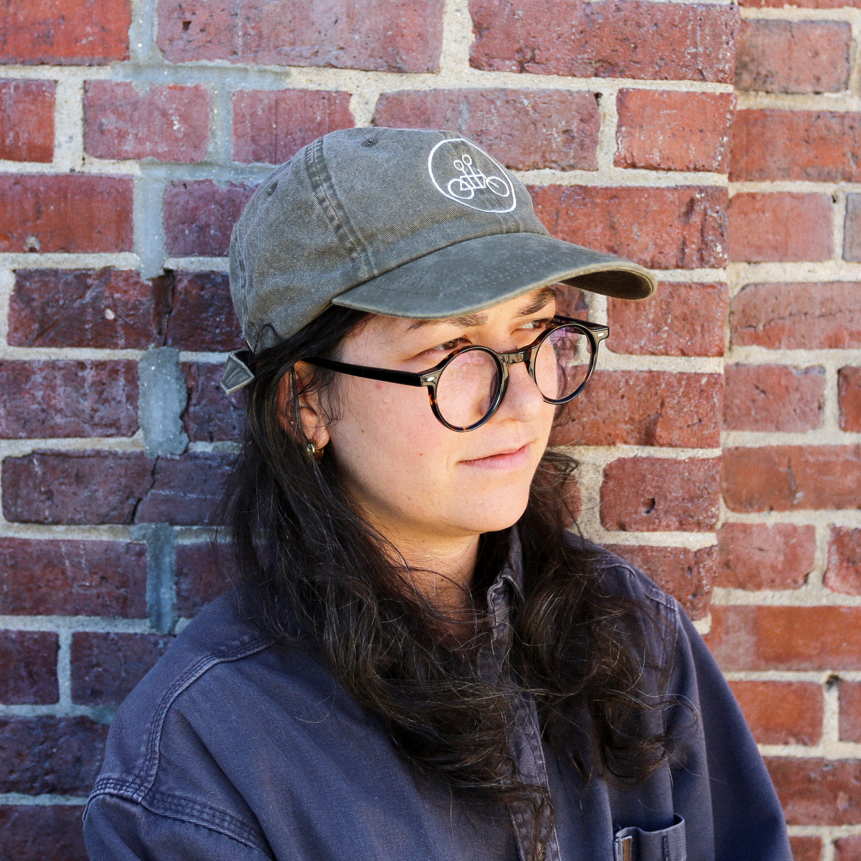 A person with long dark hair, round glasses, and a green Java Circle Logo Dad Hat by Tandem Coffee Roasters stands in front of a brick wall, wearing a blue shirt and looking slightly to the side.