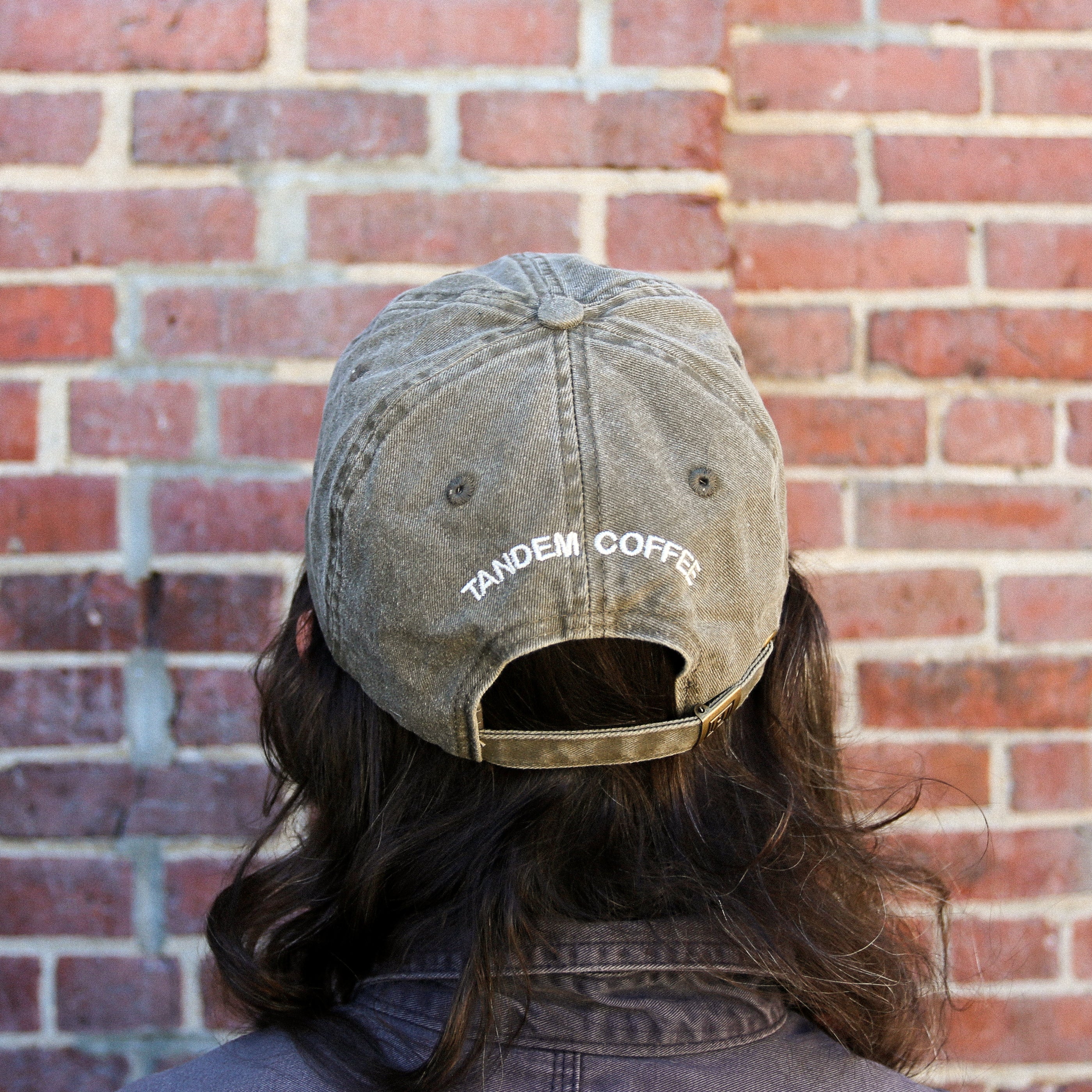 A person with long brown hair faces a brick wall, wearing a worn green Java Circle Logo Dad Hat from Tandem Coffee Roasters, featuring white TANDEM COFFEE embroidery on the back.
