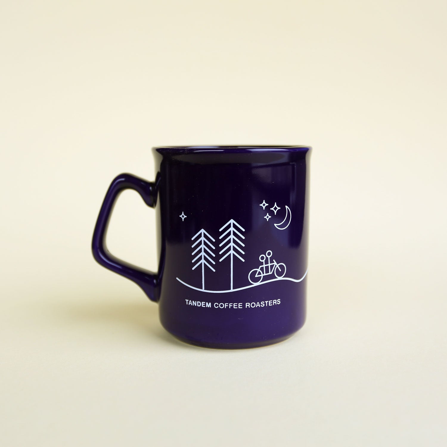 The Moon & Stars Mug by Tandem Coffee Roasters is a cobalt blue mug with a white design of two pine trees, a bicycle, a crescent moon, and stars, plus the brand name beneath. It’s shown against a plain light background.