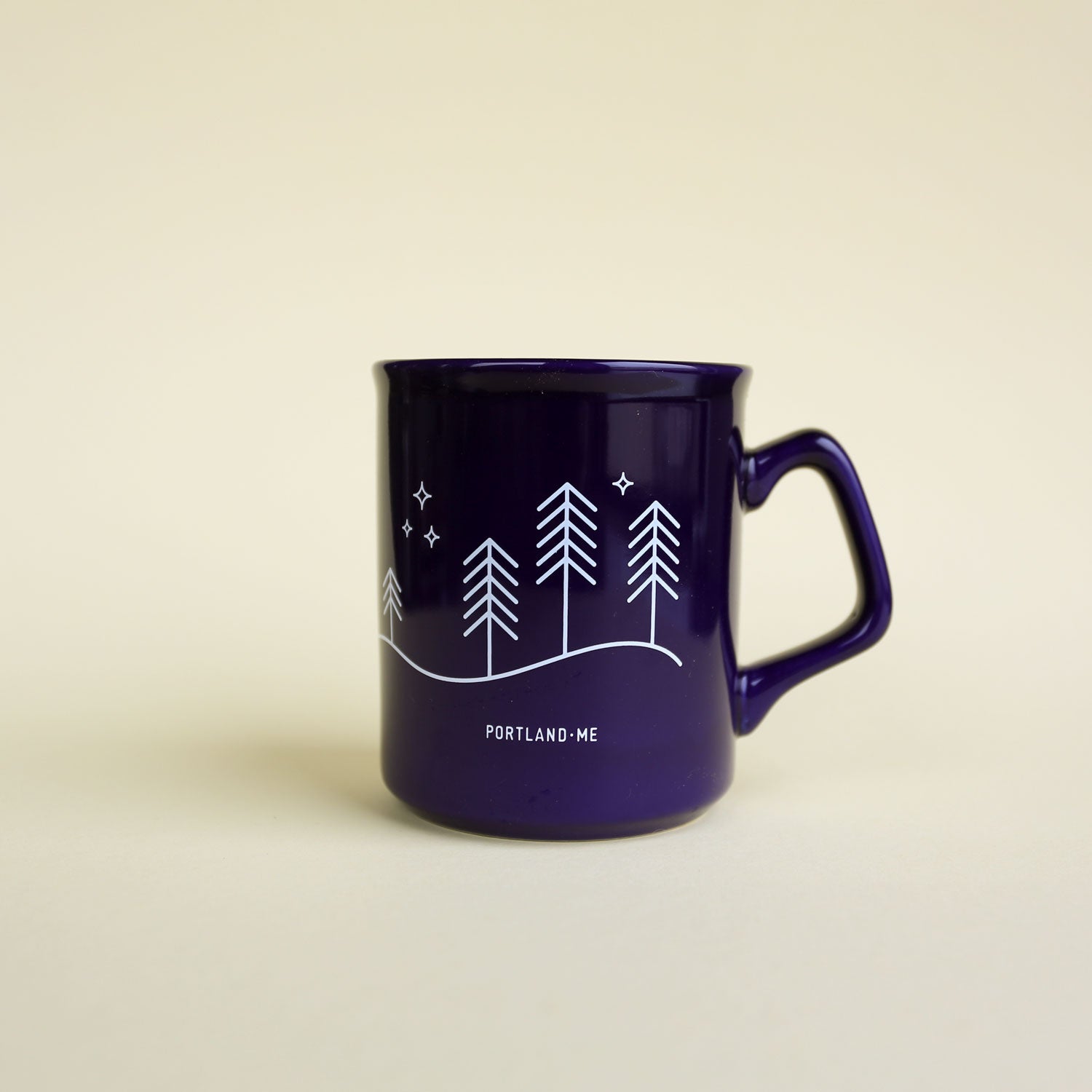 The Moon & Stars Mug by Tandem Coffee Roasters features dark purple with white line art of pine trees, hills, stars, and PORTLAND · ME beneath the design, displayed on a light beige background.