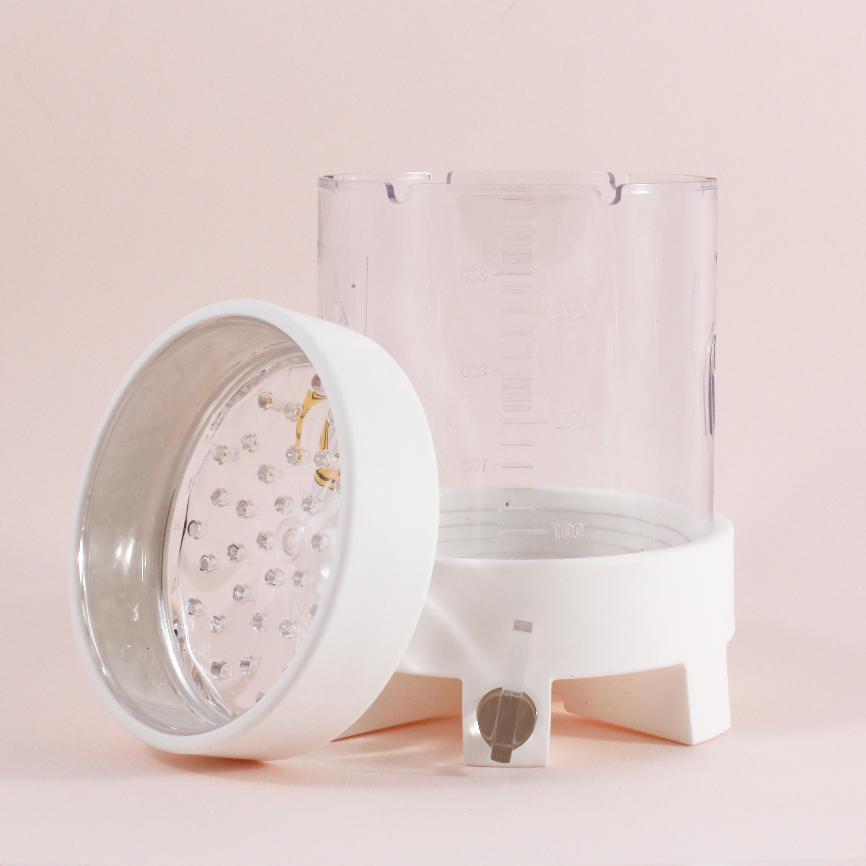 The NextLevel Pulsar Brewer by Tandem Coffee Roasters is a clear plastic manual coffee brewer with measurement markings and a white base. Its lid, featuring a metallic interior with small bumps, rests beside it against a light pink background.
