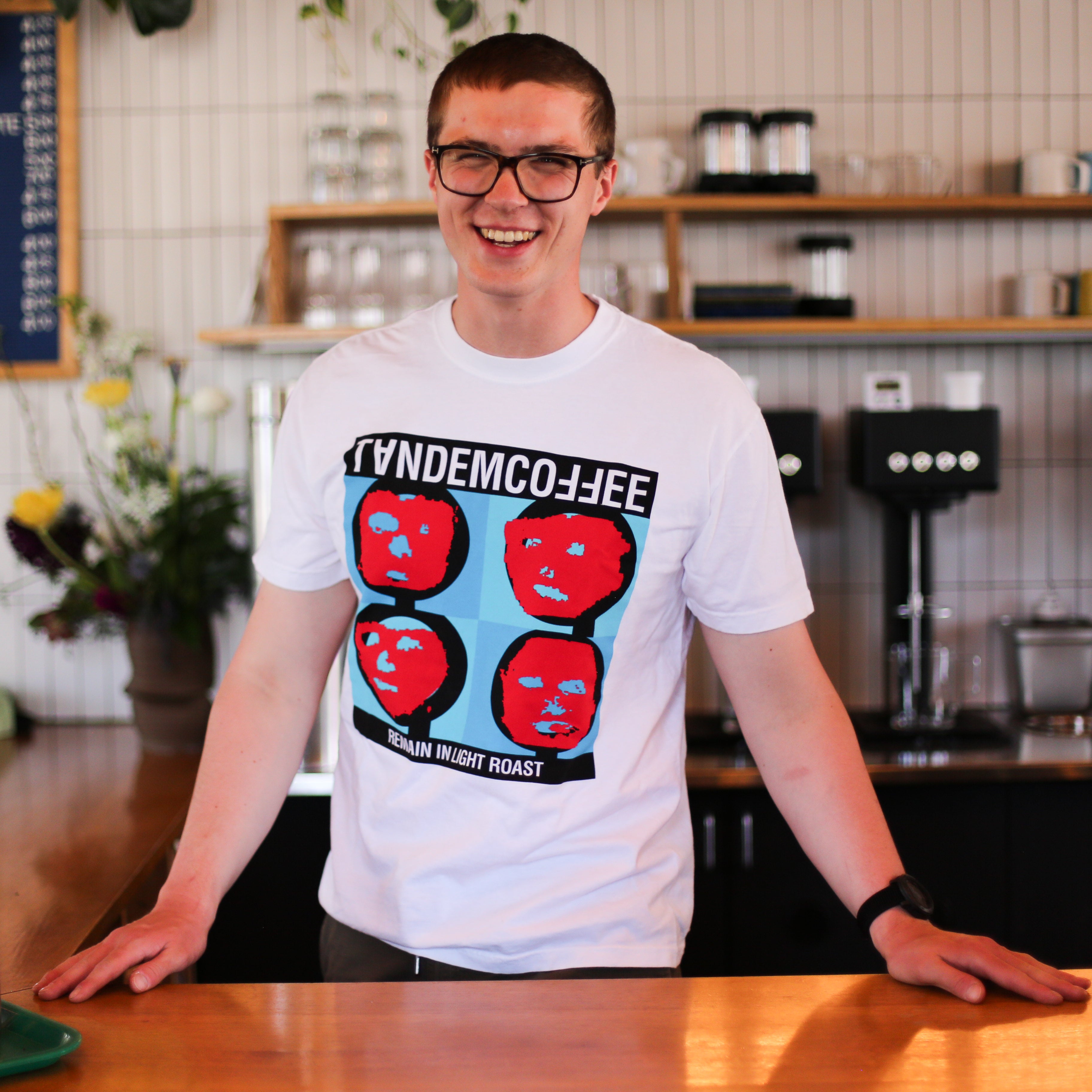 A smiling person wearing glasses and a white Remain In Light Roast T-Shirt by Tandem Coffee Roasters stands behind a wooden counter in a modern coffee shop, surrounded by shelves, plants, and coffee equipment.