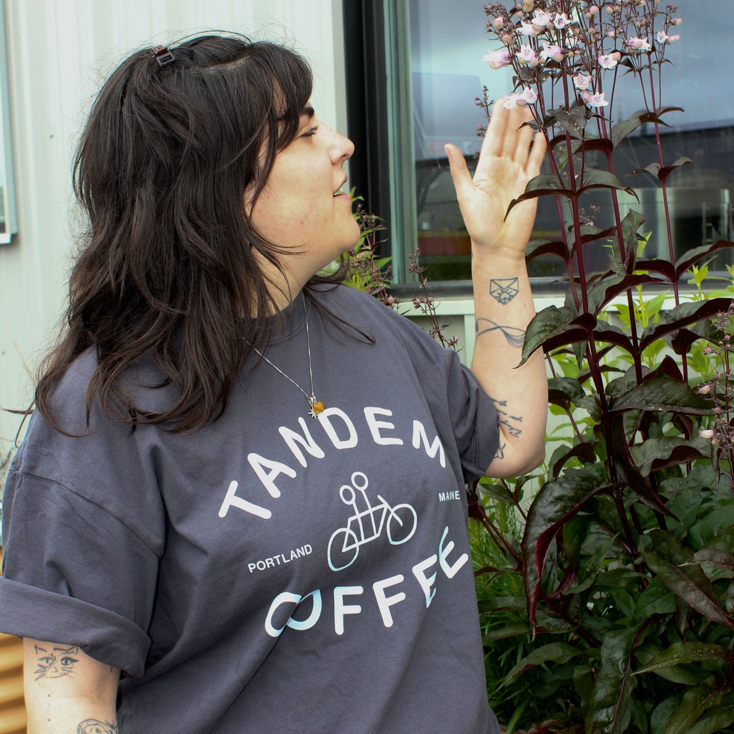 A dark-haired, tattooed person in a Tandem Coffee Roasters Graphite Logo Tee looks up at tall purple flowers, raising one hand toward them, while standing outside near a windowed building.
