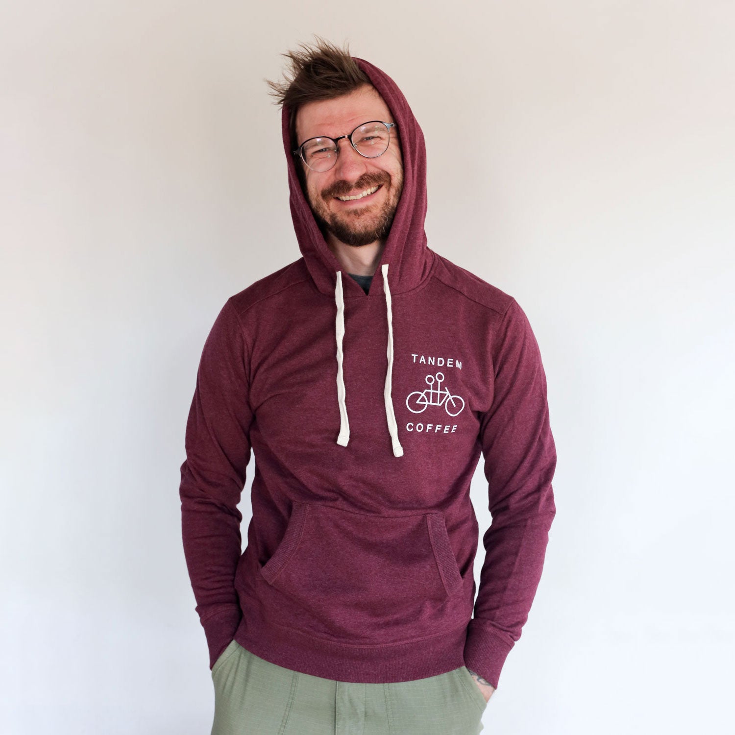 A cheerful man with glasses wearing a maroon Recycled Pullover Hoodie from Tandem Coffee Roasters, standing against a plain background, smiling broadly at the camera.