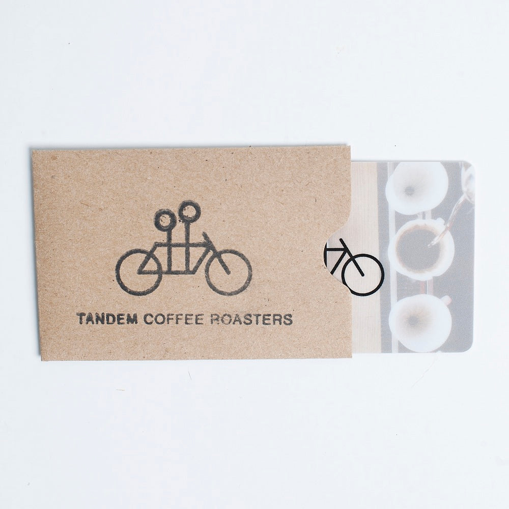 A brown sleeve with Tandem’s logo and tandem bicycle design holds a partially visible Tandem Physical Gift Card (in-store use only), perfect for treating someone to coffee and pastries.