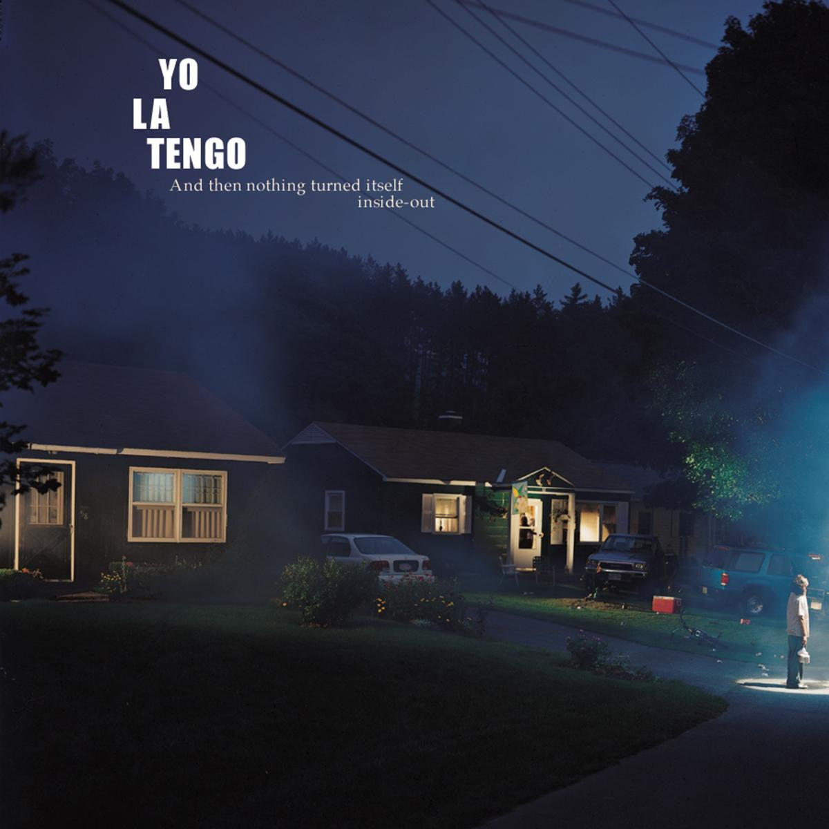 A suburban neighborhood glows at night as smoke drifts by. A figure stands in bright white light on a driveway. Text: Tandem Coffee Roasters presents Yo La Tengo - And Then Nothing Turned Itself Inside Out. Iconic indie rock album tracklist.
